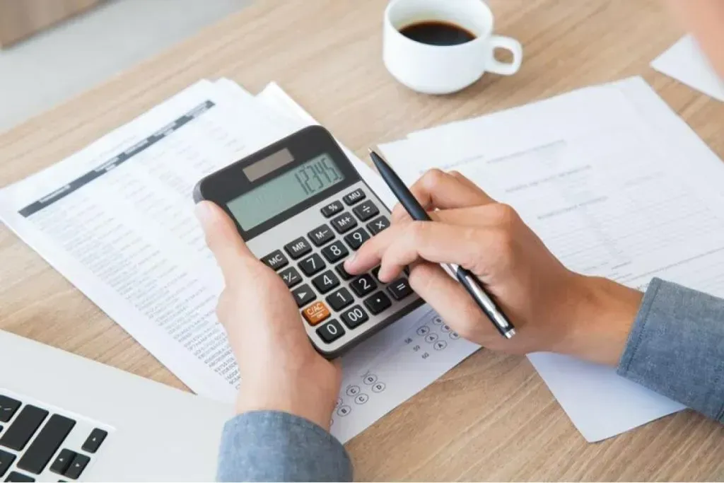 Person using a calculator and pen over printed invoices and forms on a desk